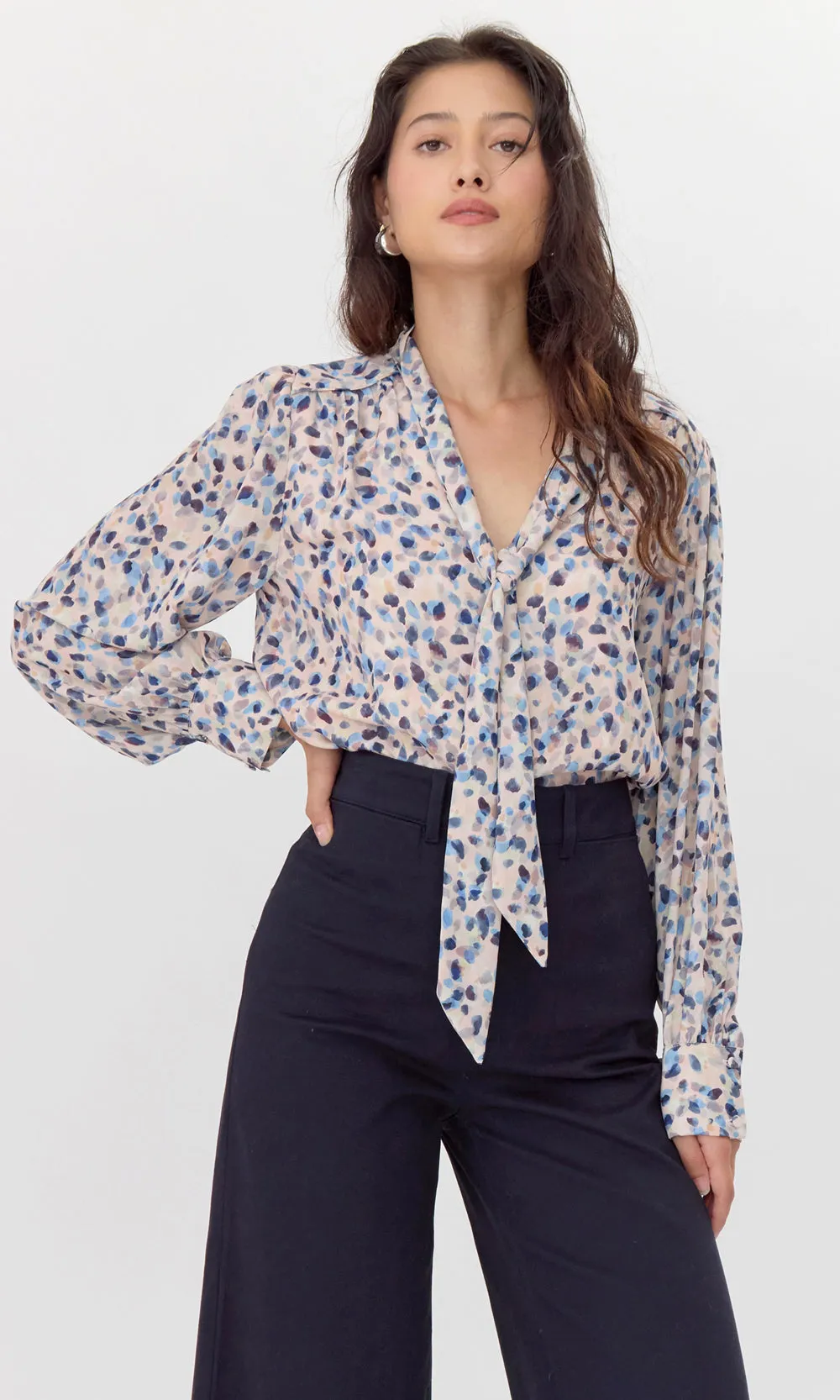 Female model wearing pink, tie-neck blouse with blue polka-dots. The blouse is tucked into her trousers.