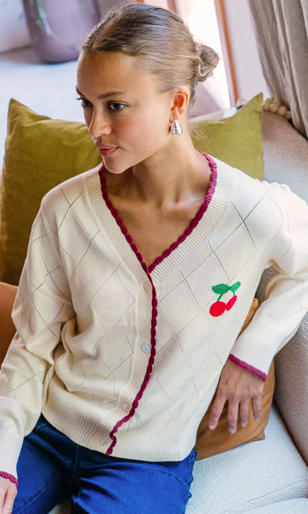 Model sits on a couch wearing a knit cardigan with a cherry design on the chest. The cardigan shows a white button closure and a red trimming along the neckline and sleeves.