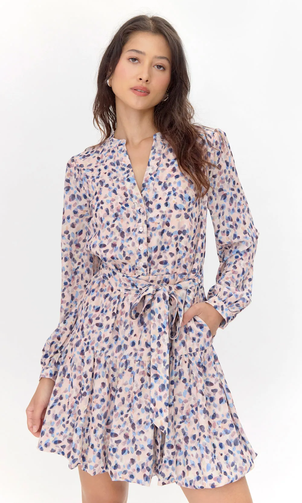 Model wearing a flowy long-sleeve dress with a pink, blue, and purple abstract print. The dress features a tiered skirt, buttoned neckline, and a matching tie belt at the waist. She poses with one hand in her pocket against a plain white background.