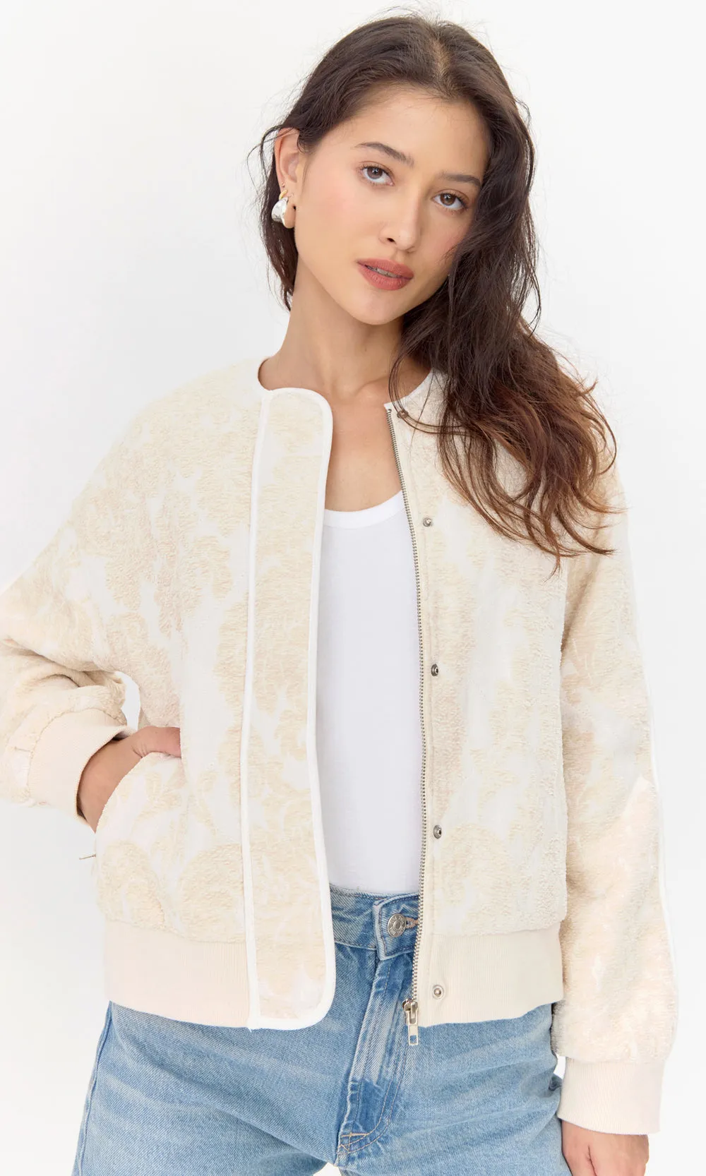 Model wears embroidered, beige jacket. She places her hand in one of the side pockets. The opened jacket shows both a zipper closure and a snap button closure.