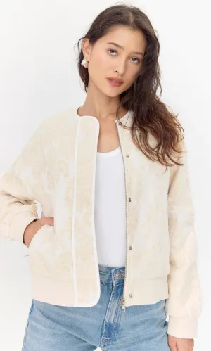 Model wears embroidered, beige jacket. She places her hand in one of the side pockets. The opened jacket shows both a zipper closure and a snap button closure.